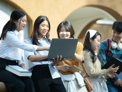 A group of college students studying together outdoors on campus, using a laptop and books in a collaborative setting.
