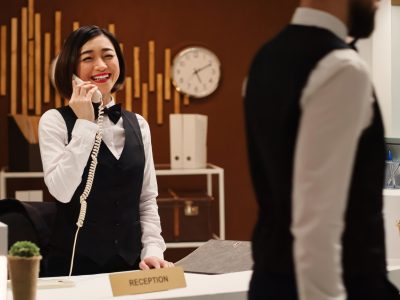 Happy smiling receptionist taking travel accommodation booking requests standing at check in desk in stylish lounge. Professional hotel employee taking phone calls while working at reception counter